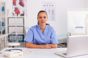 portrait-pretty-medical-nurse-smiling-camera-hospital-office-wearing-blue-uniform-healthcare-practitioner-sitting-desk-using-computer-modern-clinic-looking-monitor-medicine